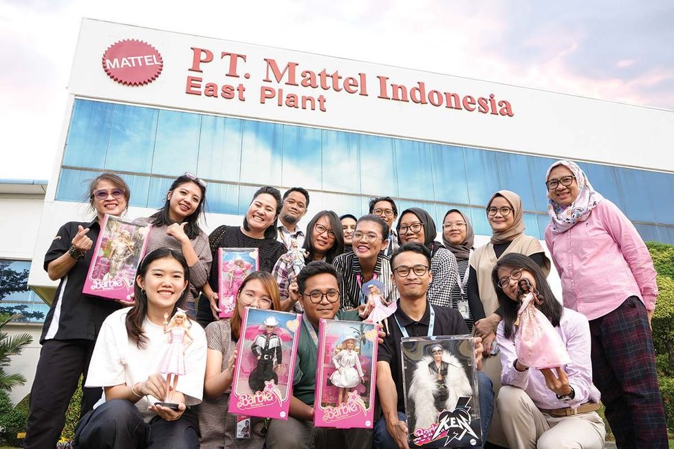 A group of women and men pose in front of a building that says, "P.T. Mattel Indonesia East Plant." Some of them hold Barbie and Ken dolls. 